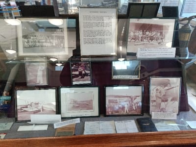 A glass-fronted, wooden display case, located in Park Hill, United States, exhibits historical artifacts primarily related to early aviation. The upper shelf features, from left to right: a large framed black and white photograph depicting a group of approximately 30 individuals, with "PLYMOUTH" visible on a background sign; a framed document titled "MICHIGAN - MAY 1928" containing dense text; a framed black and white photograph of a group of men gathered outdoors, accompanied by a descriptive label; and a dark trophy topped with a metallic eagle figurine.
The middle shelf contains three smaller framed images: a vertical black and white photograph of a single person in a light uniform; a small vertical color photograph of two individuals; and a horizontal black and white image, partially obscured by reflections, possibly showing an aerial view.
The lower shelf presents four framed black and white photographs: a horizontal image of an early biplane surrounded by several people; another horizontal image, largely obscured by reflections; a horizontal photograph showing a substantial crowd around an aircraft on an airfield; and a vertical photograph depicting two individuals interacting with a biplane on an airfield, with a descriptive label below.
The bottom surface of the display case, covered in a brown fabric, holds numerous loose historical documents, including handwritten notes, typed papers, small white cards, and a dark-covered booklet, likely a logbook or manual. Reflections of overhead lighting fixtures are visible across the glass surfaces of the display.