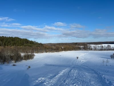 The image displays a panoramic winter landscape in Юнькаўскі сельскі Савет, Belarus, under a clear blue sky with scattered white cumulus clouds. A vast, snow-covered field dominates the foreground and midground, exhibiting numerous tracks consistent with human movement and sledding activity. In the middle distance, two dark-clad human figures are visible on the snow, one appearing to interact with a dark object, possibly a sled or toboggan. To the left, a dense evergreen forest covers a prominent hill, transitioning into a line of bare deciduous trees extending across the middle of the scene. To the right, a large body of water, completely frozen and covered in snow, extends towards the horizon. The entire scene is brightly illuminated by sunlight.