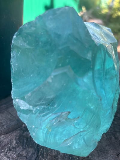 A close-up, outdoor shot focuses on a large, irregular piece of translucent, light blue glass, possibly slag glass, resting on a weathered wooden surface. The glass exhibits internal fractures and a rough, unpolished texture. In the blurred background, a dark object, possibly a pot or container, is visible to the left, and a vibrant green, out-of-focus shape, possibly foliage or a wall, is behind it. Further back, hints of sunlight filtering through greenery suggest a natural, possibly garden setting. The image does not provide any specific information about the city of Zmiiv, Ukraine, or any human presence.