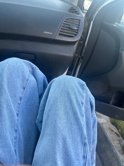 The image shows a person's legs, clad in light blue denim jeans, positioned inside a vehicle. The camera angle is from a high perspective, looking down at the person's knees and thighs. The interior of the car is visible, including a black dashboard with a textured surface and an air vent. The word "AIRBAG" is printed vertically in white on the dashboard. The car door is partially open, revealing a glimpse of an outdoor scene with grass and a concrete surface. The lighting suggests it is daytime. The overall impression is a candid, everyday moment captured from within the car, focusing on the texture of the denim and the car's interior.
