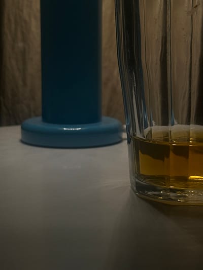 The scene depicts an indoor environment in Suginami, Japan, characterized by dim lighting. In the foreground, a clear glass with vertical fluting occupies the right side, containing an amber-colored liquid filling approximately one-quarter of its volume. To the left and slightly behind the glass, a dark teal or blue cylindrical object, possibly a lamp base, stands on a wider, flat circular base. Both objects rest on a light-colored, flat surface that spans the lower half of the image. The background consists of an out-of-focus, textured surface, light brown or beige in color, consistent with a curtain or wall. Light patterns, likely reflections, are visible on the flat surface near the glass and the blue object. No individuals, actions, or interactions are present in the scene.