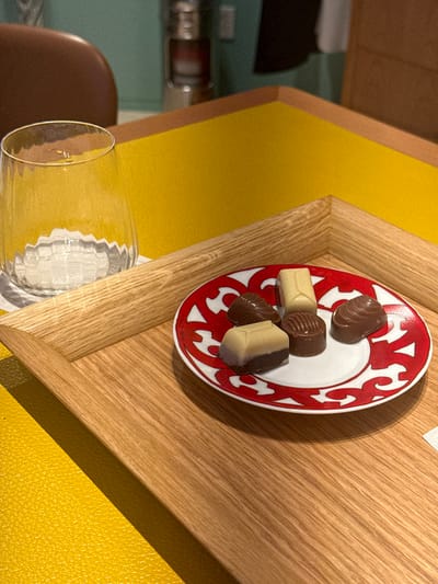 The image displays a wooden serving tray positioned on a textured yellow surface. On the tray, a small, round white ceramic plate with a distinctive red pattern and "HERMES PARIS" branding along its rim holds five assorted chocolates. These include two light-colored rectangular prisms, one dark brown conical chocolate with ridges, one small dark brown cylindrical chocolate with concentric ridges, and one dark brown shell-shaped chocolate. To the left of the plate, a clear, fluted glass containing a transparent liquid rests on a white coaster. The background features additional blurred yellow surfaces, a brown curved object likely part of a chair, and a teal-colored wall or partition, with a faint metallic object also visible. No individuals are present. The scene is an interior shot, indicative of a luxury retail or hospitality environment. Based on the branding, the location is identified as within or associated with the Hermès boutique at Plaza Andares, Zapopan, Mexico.