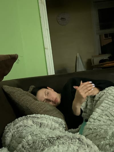 An individual is reclining on a dark brown sofa indoors, in what is identified as Dorchester, United Kingdom. The subject, a female with dark hair, lies on her left side, propped against a textured brown cushion. She wears a dark long-sleeved top. Her hands are visible near her chest, with fingernails painted a light purple or lavender shade. Her lower body is covered by a light grey, fluffy blanket. A teal-colored object, consistent in appearance with a vaping device, is partially visible protruding from under the blanket to the subject's right. The immediate background behind the subject features a light green wall and a white door frame on the left. To the right of the door frame, a dimly lit adjacent room with a light beige wall is visible. A circular, white-faced clock is mounted on this beige wall. Further to the right, a window covered by horizontal blinds is present. Below the window, a white fabric item is draped, and a dark furniture piece is partially visible. The overall lighting is dim. No other individuals are present or interacting with the subject.
