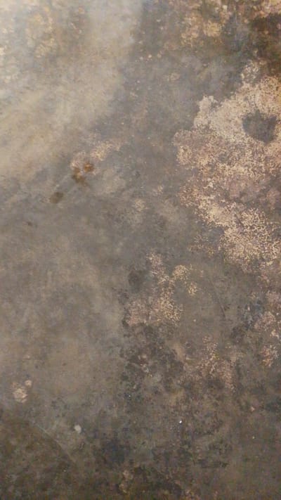 This image is an abstract close-up of a textured surface, likely a floor or wall. The surface appears to be made of concrete or a similar material, exhibiting a mottled pattern of various shades of grey, brown, and beige. There are areas with a rougher, almost granular texture, interspersed with smoother, darker patches. Some lighter, almost white specks are also visible. The lighting suggests an indoor setting, possibly with diffused natural light or artificial lighting that creates subtle shadows and highlights, emphasizing the unevenness of the surface. There is no clear indication of specific objects, people, or a particular scene being depicted. The overall impression is one of an aged or weathered surface, possibly in a utilitarian or outdoor environment. The colors and textures do not provide any explicit cues about the time of day, weather, or specific location beyond the general context of an interior or exterior surface. No text is visible in the image.