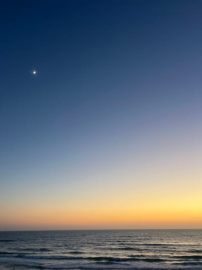 A tranquil seascape features a gradient sky transitioning from deep blue to light yellow, meeting a textured ocean surface. A faint crescent moon is visible in the darker blue portion of the sky. The water displays a pattern of subtle waves or ripples, adding depth and texture to the right edge of the image.