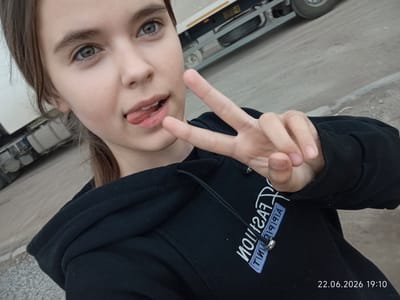 A young female with light brown hair is taking a selfie outdoors, with a truck in the background. She is sticking her tongue out slightly and holding up a peace sign with her right hand. She is wearing a black hoodie with white lettering on it that reads "Fashion" in a stylized font. The date and time stamp on the image reads "22.06.2026 19:10". The background appears to be a dirt or gravel lot with a paved surface and some construction equipment visible. The weather appears to be overcast. The location is specified as Sarytogay, Kazakhstan.