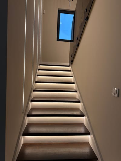 The image shows an indoor staircase illuminated with built-in LED strip lights along the wall and underneath the steps. A rectangular window is visible in the upper left corner. The staircase has dark wood panels alternating with illuminated white sections. The walls and ceiling are light beige. There is a square-shaped object on the ceiling, possibly a smoke detector or sensor. The location is unknown. There are no people in the image.