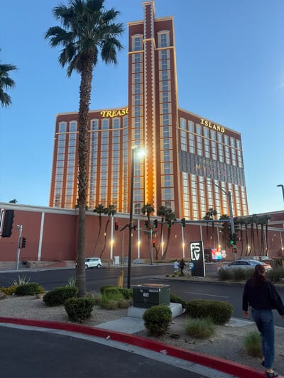 The image depicts the Treasure Island Hotel and Casino in Paradise, Nevada, USA. The tall, reddish-brown building stands prominently against a clear blue sky. The words "TREASURE ISLAND" are visible vertically on the building. A tall palm tree is in the foreground. A street runs along the right side of the frame with traffic lights, signs, landscaping, and a white vehicle. A person is walking along the sidewalk near the street.
