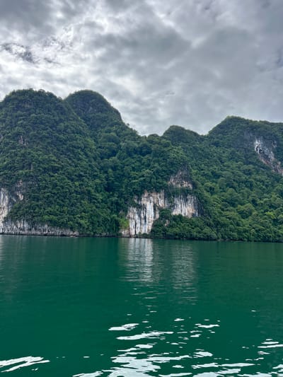 This image captures a scenic view of limestone karst formations covered in lush green vegetation, typical of the landscape found in Khlong Khian, Thailand. The formations rise steeply from the emerald green waters of a calm sea. The sky above is overcast with dramatic, textured gray clouds. The water in the foreground exhibits white reflections and ripples, indicating a gentle movement. The sheer rock faces of the karst formations show distinct vertical striations and patches of lighter colored rock, contrasting with the dense foliage that blankets their slopes and crests. The overall scene is natural and serene, with no visible human presence or artificial structures.