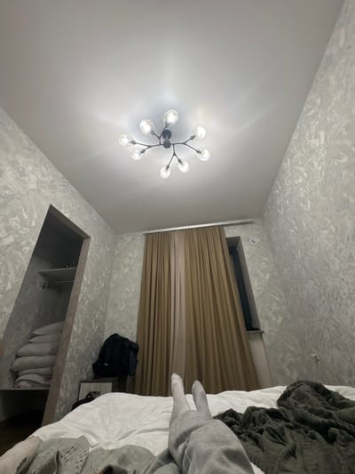 The image depicts an interior scene in what appears to be a bedroom. A modern chandelier is visible on the ceiling. A window with brown curtains is also present. A bed is on the right side of the image, covered with a white sheet and various grey and green blankets. A black backpack is near the head of the bed. Below a shelf, several items are stored, mostly white. The wall and ceiling are covered with a mottled light grey and white pattern.
The image does not provide enough information to confirm its location in Tsakhkadzor, Armenia. There are no specific landmarks or features to identify the location.