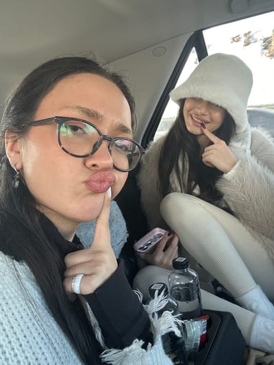 The scene depicts two females inside the passenger compartment of a vehicle, in Jraber, Armenia. The female in the foreground, positioned to the left, has dark hair, wears tortoise-shell glasses, and a light-colored knit sweater over a dark long-sleeved top. She has a white ring on her right ring finger and a small dark earring on her left ear. Her lips are pursed in a "duck face" expression, with her right index finger touching her chin. The female in the background, to the right, has long dark hair mostly covered by a white, fluffy bucket hat that obscures her eyes. She wears a light beige faux-fur coat and light-colored ribbed pants. Her left hand holds a pink smartphone with a patterned case, and her right index finger is pressed against her lower lip, which displays dark lipstick and an apparent smile. Two clear plastic water bottles with black caps and a small red and green packaged snack are visible in a cup holder or tray between the individuals. A fringed, light-colored fabric is also present in the lower center. Through the vehicle's window on the right, a bright, external environment consisting of snowy, mountainous terrain is visible.