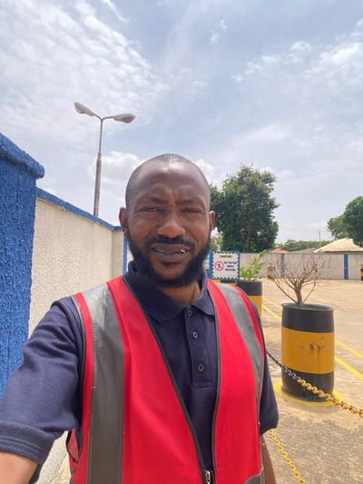 A bald Black man with a beard, wearing a navy blue collared shirt and a red and gray safety vest, takes a selfie outdoors in Kinkinau, Nigeria. He is standing in what appears to be an industrial or parking area, with a textured beige and blue wall to his left, a tall street lamp in the background, and trees visible further back. Yellow lines are painted on the concrete ground, and a yellow chain is strung between two black and yellow barrels. A sign that reads "CAUTION NO TEXTING WHILE WALKING" is mounted on a post behind the man. The sky is partly cloudy.