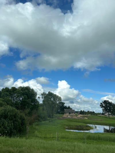 The image depicts a rural landscape under a partially cloudy sky. In the foreground, there is tall, green grass. Further back, a body of water reflects the sky. Beyond the water, a grassy field slopes upwards towards a cluster of buildings, including what appears to be a house and an unfinished structure with piles of sand and building materials. Several trees are scattered throughout the scene, with a dense line of trees forming a boundary on the left side of the image. The overall impression is one of a tranquil, agricultural area. The location is identified as Узрэцкі сельскі Савет, Belarus.