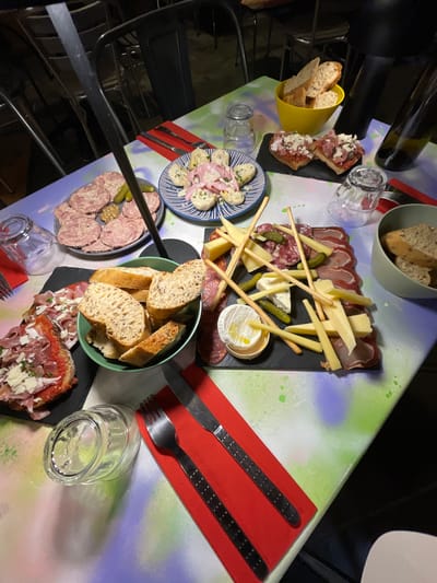 The image depicts a table laden with food, suggesting a meal in Cucuron, France. A variety of dishes are present, including several types of charcuterie, cheese, bread, and salad. Cutlery and glasses are arranged around the plates. The table is set against a vibrant, graffiti-style backdrop. No people are visible in the frame.