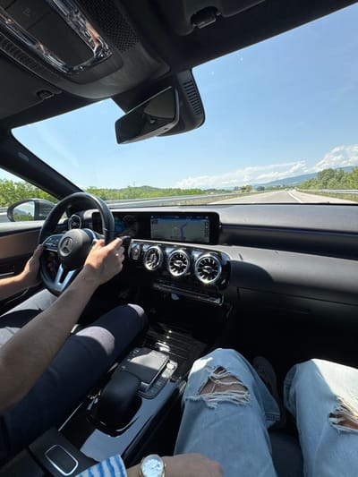 This image captures a tilted, first-person perspective from inside a modern Mercedes-Benz car, looking forward and towards the dashboard on a sunny day. The scene is oriented counter-clockwise.
Inside the car, the driver's hands are visible on the steering wheel, which prominently displays the Mercedes-Benz logo. The driver wears dark trousers. To the right, a passenger's legs, clad in light blue ripped jeans, are visible, along with a wrist wearing a silver-toned watch. The car's dashboard features a sleek design with two digital screens: an infotainment/navigation display showing a map with route details (including a "37 km" marker and a road sign icon) and a digital instrument cluster. Three circular air vents are centrally located, and the dashboard trim includes subtle ambient lighting. The center console is glossy black, equipped with various controls and a touchpad. A rearview mirror and part of the overhead console are visible in the upper left of the frame.
Outside the car, a wide, clear blue sky with a few wispy clouds is visible above. A multi-lane highway stretches into the distance, lined with white markings and guardrails. Green trees and hills or mountains form the landscape on either side of the road.
The activity depicted is driving on a highway, likely during a journey in Blajievo, Bulgaria. The clear sky and bright lighting indicate it is daytime. No specific emotions are discernible, but the setting suggests a calm and pleasant drive.
