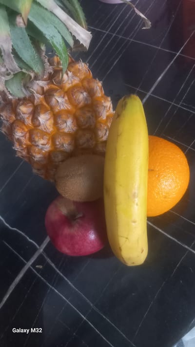 A pineapple, a banana, an orange, a kiwi, and an apple are arranged on a black tiled surface. The fruits are placed in the foreground, with the pineapple to the left, followed by the kiwi, apple, banana, and orange. The background consists of the tiled surface, which has a marble-like pattern with white lines. The overall lighting is dim, with some light coming from the top left. There is text in the bottom left corner that reads "Galaxy M32". The scene appears to be indoors, possibly on a kitchen counter or table. There are no people or animals present. The image captures a still life arrangement of fruits. The location is specified as Ынтымак, Kyrgyzstan.