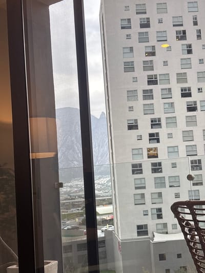 A high-rise building fills the top half of the frame, reflected in a large window. Many square windows dot the building's facade. Below, a grey mountain range appears, also reflected in the window. A dark window frame spans the middle, and interior walls and furniture are visible at the bottom, indicating an indoor setting. A hanging woven object is partially visible in the upper right corner.