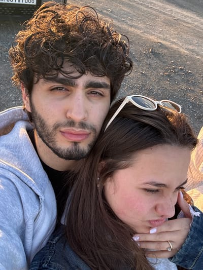 The image shows two people lying down outside at dusk. The person on the left has curly dark hair, a beard, and is wearing a gray hoodie. The person on the right has long brown hair, white sunglasses resting on her head, and is wearing a denim jacket. Her eyes are closed, and her hand is visible with a ring on her finger. They appear to be lying on a rough, gray surface, possibly asphalt or gravel. The angle of the photo suggests they are lying down looking up. The bottom left corner of the image shows a license plate with the characters "008 MT 90." The location cannot be determined as Hrazdan, Armenia based on the visual information provided.