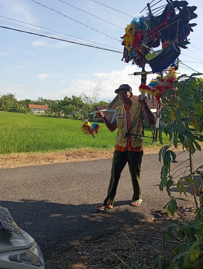 A man in Waung, Indonesia, stands on an asphalt road next to a rice paddy. He wears a tan jacket over an orange shirt, green pants, a cap, and sandals. He carries a contraption with colorful, horse-like puppets. He holds one puppet up in his hand, displaying it. Trees and bushes line the road, and power lines are visible in the sky. A portion of a car is visible in the lower right corner.