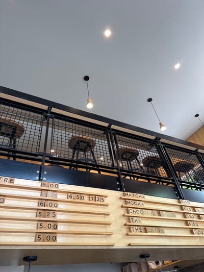 The image captures an interior view of a business in Chelsea, Canada, focusing on architectural and decorative elements. The left side of the frame features a white wall with embedded circular lights, some of which have small, black cable-like structures extending from them. In the center of the image, there is a dark metal structure with a wire mesh, resembling a staircase or a decorative railing. Behind the mesh, there are wooden chairs. To the right, a wooden menu board displays a list of items with their prices. The items listed are: MATCHA, MOCHA, CHOCOLAT CHAUD, THE, and THE LATTE, each with its corresponding price in dollars. The menu is mounted on a wall, next to a dark cylindrical object attached to a black rod. The camera angle is tilted upwards, creating a skewed perspective of the scene.