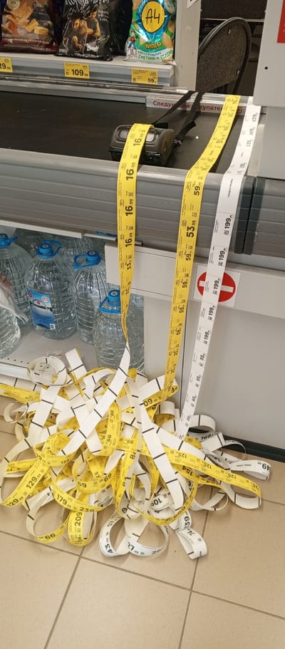 In Zhirnovsk, Russia, a retail checkout area displays a cluttered pile of printed receipt tape on the tiled floor. The receipts are yellow and white, with black text and lines indicating prices and potentially other product information. A receipt printer is visible on the checkout counter, with two long receipt streamers extending from it, one yellow and one white, partially unspooled. These streamers are intertwined with the pile on the floor. Behind the counter, shelves are stocked with large plastic bottles of water. The environment suggests a commercial setting, possibly a grocery store or supermarket, with the floor tiles and shelving units being characteristic of such a location. There are no people visible in the image.