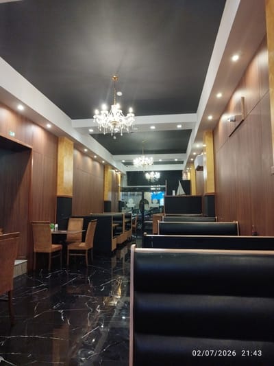 The image depicts the interior of a restaurant in Fgura, Malta. The scene is dimly lit, with a focus on the dining area. Dark, glossy marble flooring reflects the ambient light. The walls are paneled with dark wood, accented by gold-colored columns. The ceiling is black, with recessed lighting and two ornate chandeliers hanging down.
In the foreground, several rows of black upholstered booth seating are visible, facing towards the camera. To the left, a small dining table is set with two brown chairs. Beyond that, more booth seating and tables are arranged in the restaurant. In the background, through large glass windows, a street view is visible, showing parked cars. A date and time stamp of "02/07/2026 21:43" is present in the bottom right corner of the image. There are no people visible in the photograph.