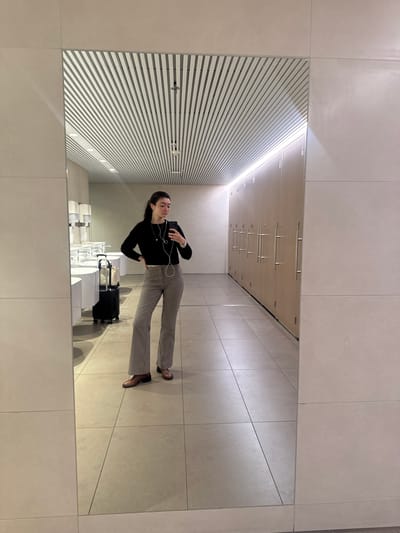 A mirror selfie captures a young woman standing in what appears to be a modern public restroom or washroom. The image is taken from a slightly elevated perspective, showcasing the interior reflected in a large, clean mirror that covers much of the wall.
The woman is the central subject, reflected upright in the mirror. She has dark hair, pulled back, and is wearing a long-sleeved black top and checked (houndstooth or plaid) trousers. Brown shoes complete her outfit. She holds a smartphone in her right hand, angled towards herself, indicating she is taking the photo. White wired earphones are visible, connected to her phone and resting around her neck. Her expression is neutral, with her gaze directed towards the phone. Next to her, on the light-colored tiled floor, is a black, hard-shell rolling suitcase, suggesting she is traveling.
The reflected background reveals a contemporary restroom design. Multiple white, rectangular sinks are visible, along with wall-mounted dispensers. Above these, there is a prominent slatted wall feature, possibly part of the ceiling or a decorative element, with integrated linear lighting that emits a bright glow. Further back, above the slatted design, light-colored wooden cabinets or panels with horizontal silver handles are reflected. The floor and visible wall sections outside the mirror are tiled in a uniform, light grey or beige color, contributing to a clean and minimalist aesthetic.
The setting, with its modern design and the presence of luggage, strongly suggests a public space, possibly an airport or a train station in Aalsmeerderbrug, Netherlands. The lighting is artificial, giving no indication of the time of day or outdoor weather. No visible text is present in the image.
