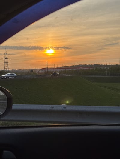 The image depicts a sunset viewed from inside a vehicle, possibly on a highway near Poroshkino, Russia. The sun is setting towards the left side of the frame, casting a warm, golden light on the sky. In the foreground, there is a glimpse of the vehicle's interior, including the side mirror and part of the dashboard. Outside the vehicle, there is a grassy area bordering the road. Power lines and towers are also visible, silhouetted against the setting sun. Further in the distance, faint outlines of buildings and infrastructure suggest a suburban or peri-urban environment. Cars are visible on the road.