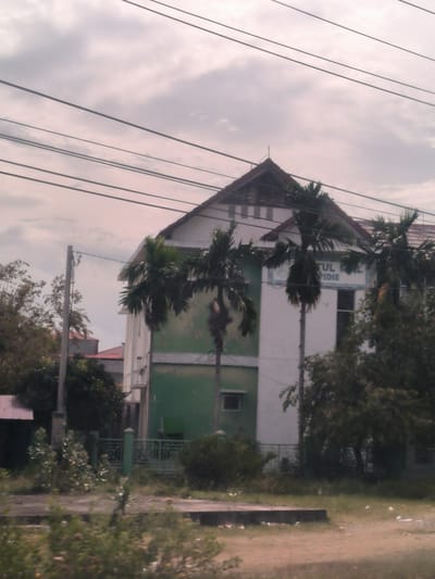 This daytime image captures an outdoor scene in Lampeudeu Baroh, Indonesia, featuring a multi-story building, likely an institutional or school facility, under an overcast sky.

The central subject is a white building with a prominent, dark-tiled pitched roof. The lower part of its facade is painted a distinct green, contrasting with the white upper section. Several tall palm trees are positioned in front of and around the building, partially obscuring its details. Other smaller trees and bushes are visible in the mid-ground and background.

In the immediate foreground, there is an unkempt area of sparse vegetation and dirt, traversed by a low concrete curb or platform. A green metal fence acts as a boundary in front of the building. To the left, a utility pole rises, from which multiple black power lines stretch horizontally across the upper part of the frame.

The sky is cloudy and grey, indicating an overcast day with soft, diffused lighting and no harsh shadows. No specific activity or people are visible in the static scene.

Visible text on the building's facade, positioned between two palm trees, appears in blue lettering. Fragments of the text are legible, reading "FAKULTAS" (partially visible on the left), "UL ML" on the upper line, and "PIDIE" on the line below.