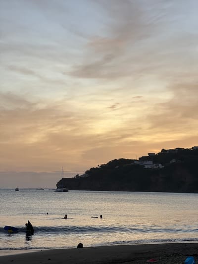 The image captures a serene evening at a beach in San Juan del Sur, Nicaragua. The sky is painted with soft hues of orange and yellow, transitioning into a pale blue, indicating either sunrise or sunset. Wispy clouds are scattered across the horizon. The calm ocean water reflects the sky's colors, with gentle waves lapping at the dark sand beach.
In the foreground, a few individuals are visible in the water. Two people are near the shore, one appearing to be standing and interacting with a blue floatation device, while another person is also in shallow water. Further out, several other figures are present, some swimming and one lying on a floatation device.
A sailboat with a white hull and a tall mast is anchored in the bay, its sail down. Another smaller boat is visible further in the distance. The background features a lush, dark green hillside dotted with white buildings, suggesting houses or villas. The overall atmosphere is peaceful and tranquil.