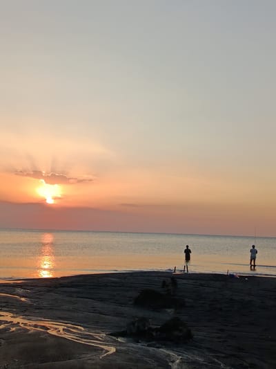 The image depicts a serene beach scene at sunset in Jangkar, Indonesia. The sun, partially obscured by clouds, casts a warm, orange glow across the sky, with rays of light fanning out above. The tranquil ocean reflects the vibrant colors of the sunset, creating a shimmering path on the water.
Two figures are visible in the shallow water, engaged in fishing. One person, silhouetted against the sunset, stands in the water holding a fishing rod. To their right, another individual, also with a fishing rod, is partially submerged in the water. The foreground features dark, wet sand with patterns formed by receding water, suggesting it might be low tide. Scattered dark objects, possibly driftwood or rocks, are present on the sand.
The overall mood of the image is peaceful and tranquil, capturing the quiet beauty of a coastal evening. The soft light and calm waters evoke a sense of relaxation and the simple pleasure of enjoying nature. There is no visible text in the image.