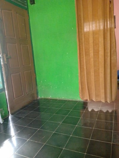 The image depicts an interior scene in Sukamanah, Indonesia, focusing on a corner where a brown wooden door meets a bright green wall. The door, slightly ajar, reveals a sliver of a lighter-colored interior and has a silver and brass door handle. To the right of the green wall, a light brown pleated curtain hangs, obscuring whatever lies beyond. The floor is tiled with large, dark, square tiles that are highly reflective, showing streaks of light and the faint outlines of the pleated curtain. Some small white speckles are visible on the green wall and on the floor tiles. The lighting suggests it is daytime, with natural light entering from the left, casting a bright glare on the floor near the door. The scene is static and there is no visible text. The overall impression is of a simple, possibly residential, interior space.