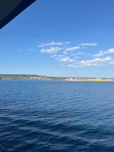 The image shows a body of water, likely the sea, in the foreground. The water is a deep blue and has small ripples on its surface, indicating a calm or lightly breezy day. In the background, a coastal town is visible, nestled along a green, hilly coastline. The town appears to be composed of numerous white buildings with red roofs, characteristic of Mediterranean architecture. Further to the right, a large, yellow structure, possibly a ferry or cargo ship, is docked at a port. The sky above is a clear, vibrant blue, with scattered white clouds. A portion of a dark blue structure, possibly part of a boat or building, is visible in the top left corner, framing the scene. This view is consistent with the coastal area of the Municipal Unit of Kastro - Kyllini, Greece, known for its picturesque towns and proximity to the sea. There are no visible people in the image.