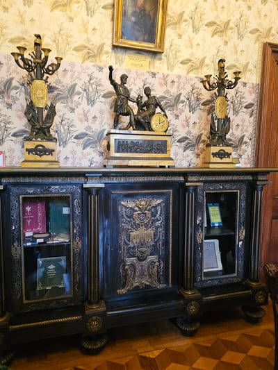 The image shows an interior view, likely within a historical building or museum in Алупкинский городской совет, Ukraine. The scene features an antique wooden cabinet with intricate carvings and decorative elements. The cabinet has shelves containing books and other artifacts. Adjacent to the cabinet, a wall covered in patterned wallpaper holds mounted clocks and statues. The overall environment suggests a room of significant historical or artistic value.
