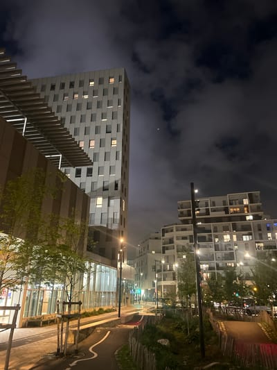 The image depicts a nighttime urban scene in Ivry-sur-Seine, France. Two modern residential buildings are visible, one with a white grid-like facade and the other with a more conventional design. A street with trees and foliage lines the edge of the frame, leading to a commercial area. A crescent moon is visible in the cloudy sky. The lighting is artificial, emanating from street lamps and illuminated windows in the buildings.
