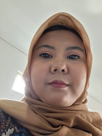A person wearing a light brown hijab looks directly at the camera from a low angle. The person has fair skin, dark eyebrows, and dark eyes. Lips appear to have light red tint. Part of a white ceiling is visible in the background, along with a bright square shape. The individual's clothing features a dark blue and white patterned design.
