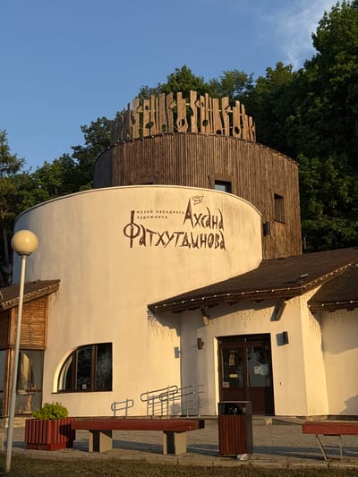 The image shows the exterior of the Museum of Folk Artist Fatkhutdinova in Krasny Klyuch, Russia. The building has a unique architectural style, featuring a rounded tower-like structure with wooden planking details at the top. A sign on the side of the tower displays the name of the museum in a decorative font. Part of the building has a white wall that connects to a rectangular building. The exterior includes windows, doors, a bench, bicycle rack, and decorative lighting. Green trees and a clear blue sky form the backdrop of the scene.