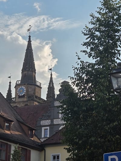 The image captures a view of the Ansbach Town Hall (Rathaus), a prominent historical building in the city of Ansbach, Germany. The focus is on the tower and parts of the surrounding architecture. The tower, constructed of stone, features intricate stonework and ornate detailing, particularly on its spire. A large clock face, with a blue and white patterned dial, is visible on the tower. Two smaller spires, adorned with flags, flank the main tower.
In the foreground, to the right, is a large, leafy tree, its branches obscuring parts of the sky and the building. The leaves are a rich green, suggesting it's likely a temperate season. Peeking out from behind the tree on the far right is a street lamp with a glass lantern.
The background consists of a bright, partly cloudy blue sky. The lower section of the image reveals the red-tiled roofs of adjacent buildings, with visible windows and architectural embellishments on one of the structures. The scene depicts a picturesque urban landscape, likely taken from a street level perspective, looking upwards towards the historic town hall. No people or specific actions are depicted.