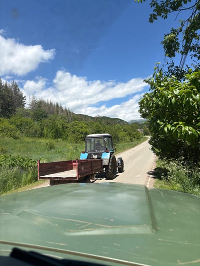 In Jermuk, Armenia, a blue tractor with a red trailer is driving on a paved road. The tractor is moving away from the viewer. The trailer is empty. The road is surrounded by green grass and trees. The sky is blue with some white clouds. The view is from inside another vehicle, with its green hood visible in the foreground.