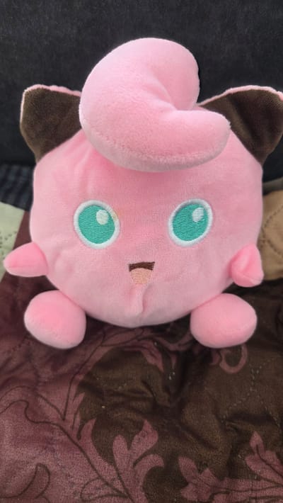 A pink plush toy with large, teal eyes and small brown ears rests on a patterned brown and pink fabric. The toy appears to be a character with an elephantine trunk-like feature. The environment appears to be indoors, possibly on bedding or furniture. There is no indication of a location in Sannikovo, Russia.
