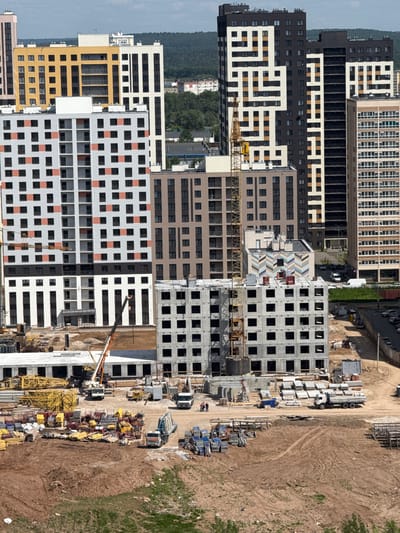 The image shows a large construction site in Shul'gino, Russia, with several tall residential buildings in various stages of completion. In the foreground, the foundation and lower floors of a concrete building are visible, with scaffolding and construction materials scattered around. A large yellow crane is actively working at the site, lifting materials to higher floors. Various trucks and other construction vehicles are also present. In the background, several completed apartment buildings are visible, indicating this is a new development area. The overall scene is one of ongoing urban development.