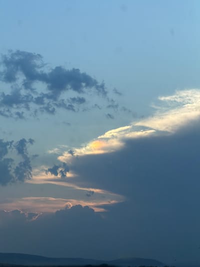 This is a photograph of the sky above Drazhevo, Bulgaria, taken at dusk. The sky is a gradient of pale blue at the top, transitioning to darker blues and grays towards the horizon. Several clouds are visible, some appearing as wispy formations and others as darker, more dense masses. Notably, a segment of cloud near the center exhibits iridescence, displaying faint rainbow colors. In the distance, a silhouetted landscape of rolling hills and distant trees marks the horizon line. The overall impression is that of a serene, twilight sky.