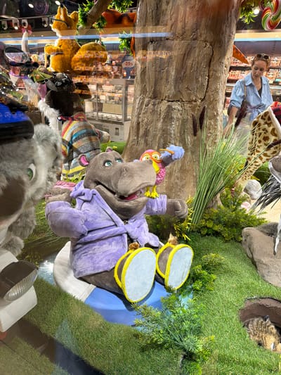 In Sokilnyky, Ukraine, a store window display features plush toys in a forest-like setting. A large, grey hippopotamus plush toy, wearing a purple robe, sits on a blue cushion with yellow-trimmed feet. To its left, a grey plush creature with green eyes is partially visible. Behind the hippopotamus, a plush owl with pink and yellow feathers is perched, holding a blue object, possibly a toothbrush, in its beak. A giraffe plush's neck and head are visible to the right, and a furry brown creature peeks out from a hole in the ground on the far right. The display includes artificial grass, a tree trunk, and dried reeds. In the background, through the window, a woman in a light blue shirt is browsing items on shelves. The scene appears to be a whimsical display likely intended for children.