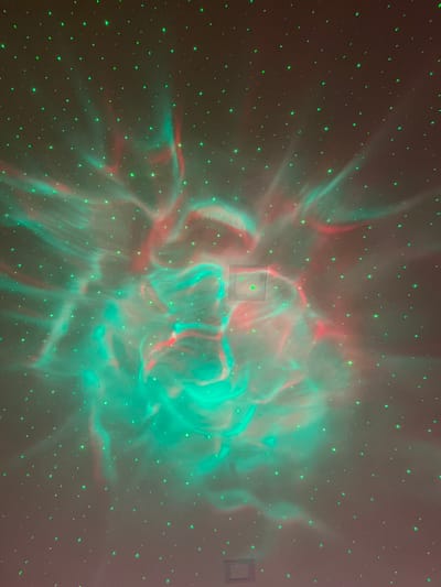 The image displays an interior wall or ceiling in Pedrouços, Portugal, illuminated by a light projector. The surface is a uniform, light-toned material, likely painted plaster. The projection consists of a dense field of small, distinct green laser points scattered across the entire visible area, resembling stars. Overlapping these points are dynamic, swirling, nebulous light formations primarily in vibrant green and reddish-pink hues, intermingling to create fluid, cloud-like patterns. These nebulous patterns are more concentrated in the central and lower portions of the frame. A rectangular electrical fixture, possibly a switch or outlet cover, is visible in the upper-right central area, partially illuminated, with a single green indicator light. Another darker, rectangular fixture is present near the bottom edge, centrally located. No individuals or direct human actions are depicted. The environment is dimly lit, with the projected lights serving as the primary visual element.