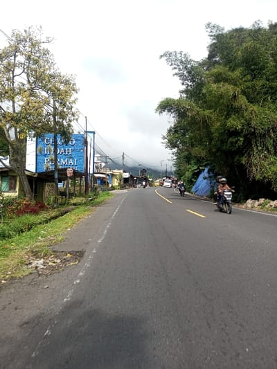 The image depicts a daytime view of an asphalt road curving slightly right, under an overcast sky, in a location identified as Saing Ta Mawt Lay, Myanmar.
On the left side of the road, a large blue sign with white text, partially obscured by a tree with light green foliage, reads "CICO INDAH PERMAI Resort & Hotel." Behind this sign and further along the left roadside are several modest, single-story structures, including a building with light-colored walls, with overhead power lines. The immediate left foreground shows a grassy verge and a small drainage ditch.
The right side of the road features a steep embankment densely covered in green bamboo and other tropical vegetation, with a blue tarp visible within the foliage. The two-lane road has a faded white line marking its right edge and a prominent yellow center line that transitions to a double yellow line further into the distance.
Traffic primarily consists of motorcycles moving away from the viewer. A dark motorcycle in the right lane carries two helmeted riders in the foreground. Further down the road, at least two additional motorcycles and a white van are visible. In the background, distant, dark, forested mountains are partially obscured by atmospheric haze or clouds. The sky is uniformly white and overcast.