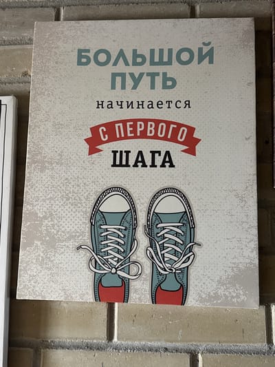 The image shows a poster with a motivational message in Russian. The phrase "Большой путь начинается с первого шага" translates to "A long journey begins with a single step." The poster features a pair of illustrated sneakers. The background has a textured, speckled appearance. The poster is hung on a brick wall. The location in the city of Zamankul, Russia, cannot be confirmed from the image.