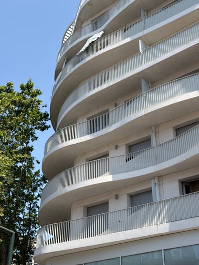 The image captures a modern, multi-story building with a curved facade, featuring numerous balconies with light-colored railings. The building is constructed from concrete and appears to be residential or commercial. A lush green tree with dense foliage is visible on the left side of the frame, partially obscuring the building. The sky above is clear and blue, indicating a sunny day. The building's design is characterized by its smooth, flowing lines and repetitive balcony structures. In the foreground, a partial view of a metallic structure, possibly part of a fence or gate, is visible at the bottom left corner. The scene is set in Courbevoie, France, as indicated by the metadata. There are no visible people in the image. The overall impression is one of contemporary urban architecture set against a natural element of a large tree.