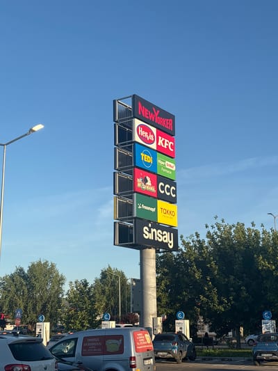 The image shows a tall, multi-panel signpost advertising various businesses in Giurgiu, Romania. The signpost features prominent logos for New Yorker, Hervis, KFC, Tedi, Hiper Dr.Max Farmacie, XXXLutz, CCC, Fressnapf, Takko Fashion, and Sinsay. The background consists of a clear blue sky and lush green trees. In the foreground, several cars are parked, and a street lamp is visible on the left. The overall scene depicts a commercial area, likely a shopping center or retail park, on a sunny day.