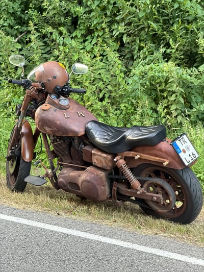 The image shows a rusty custom motorcycle parked on the side of a road, likely in Mensdorf, Luxembourg. The motorcycle has a brown helmet with a clear visor placed on its handlebars. The bike features a black seat with flame-like stitching, and a license plate with the letters "TR" and numbers "L 16". The background consists of lush green foliage. The road surface is asphalt with a white line marking its edge. The motorcycle is the primary subject, positioned prominently in the frame. No people are visible in the image.