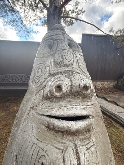 A large, grey, carved wooden statue of a face is situated outdoors, likely in a garden or park. The statue's face features circular carvings for eyes and a deep, dark opening for the mouth. A small tree is partially visible on the left side of the statue, while a dark wooden structure is in the background, possibly a building or fence. The ground is covered with dry grass and other vegetation. The sky above is partly cloudy. No people are visible in the image. There is no information within the image to confirm that the image was taken in Sukhobuzimskoye, Russia.